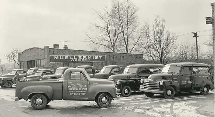 The Muellermist yard around 1950: a brick office building with a
                  painted MUELLERMIST OFFICE sign and a fleet of hand-lettered panel trucks